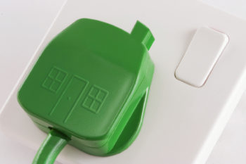 A green United Kingdom 3 Pin plug as a symbolic Green home, plugged into a white wall socket.