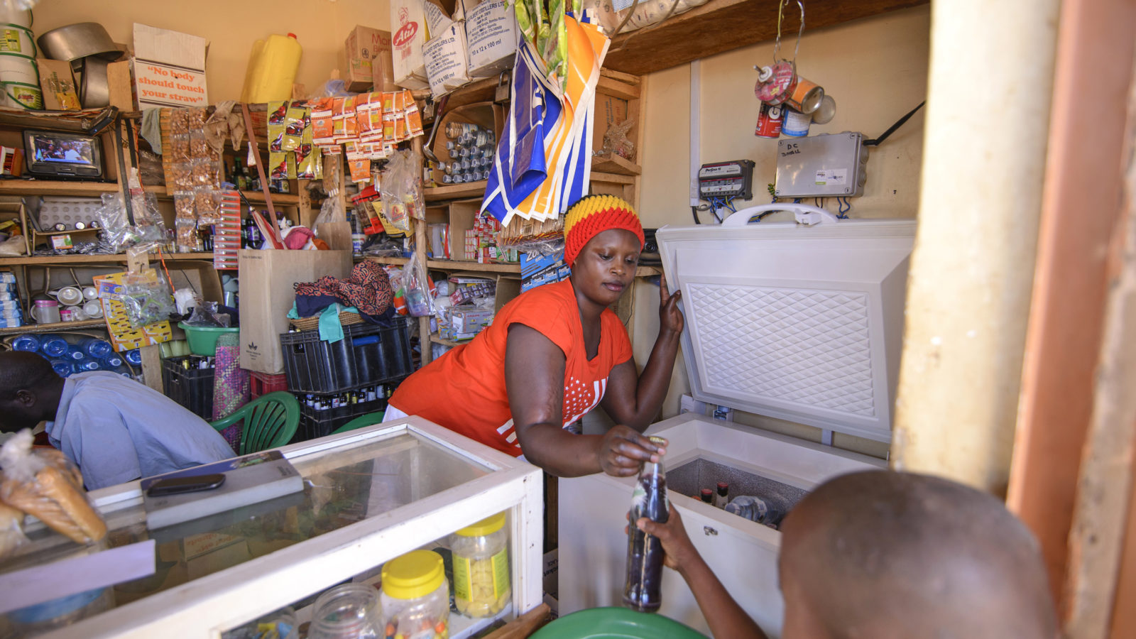 A black woman in her store, leans over the counter to hand a child a cool bottle of soda from her fridge