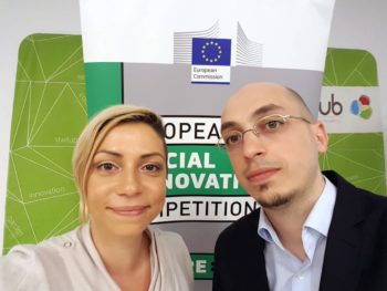 Two people stand in front of a sign for the European Social Innovation Competition