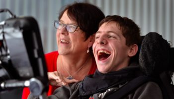 A young man in a wheelchair smiles with a woman in red next to him