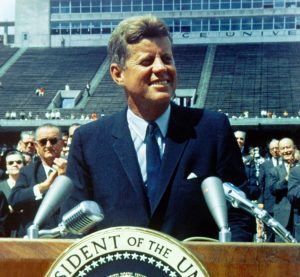 President John F Kennedy standing on a podium looking off and smiling to the right
