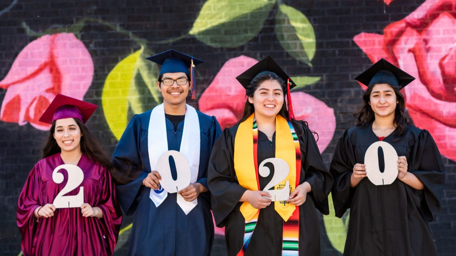 Four graduates stand in different colour robes holding the numbers for 2020. There is a floral pattern behind them with big pink roses.