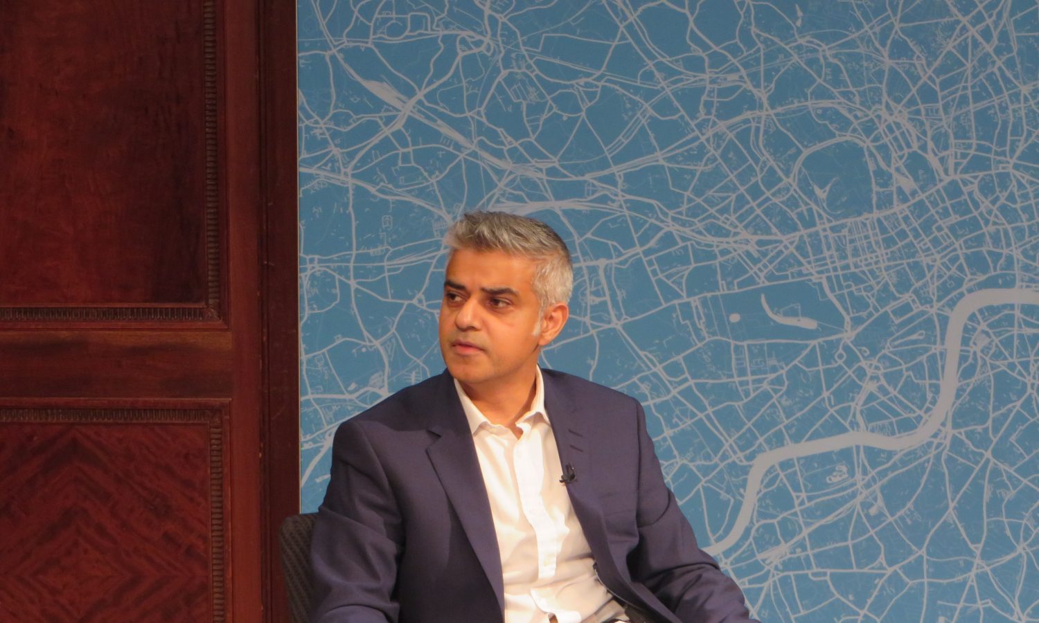 London Mayor Sadiq Khan sits in the centre of the pic looking to the left. The background is a wall with blue wallpaper outlining the city of London.