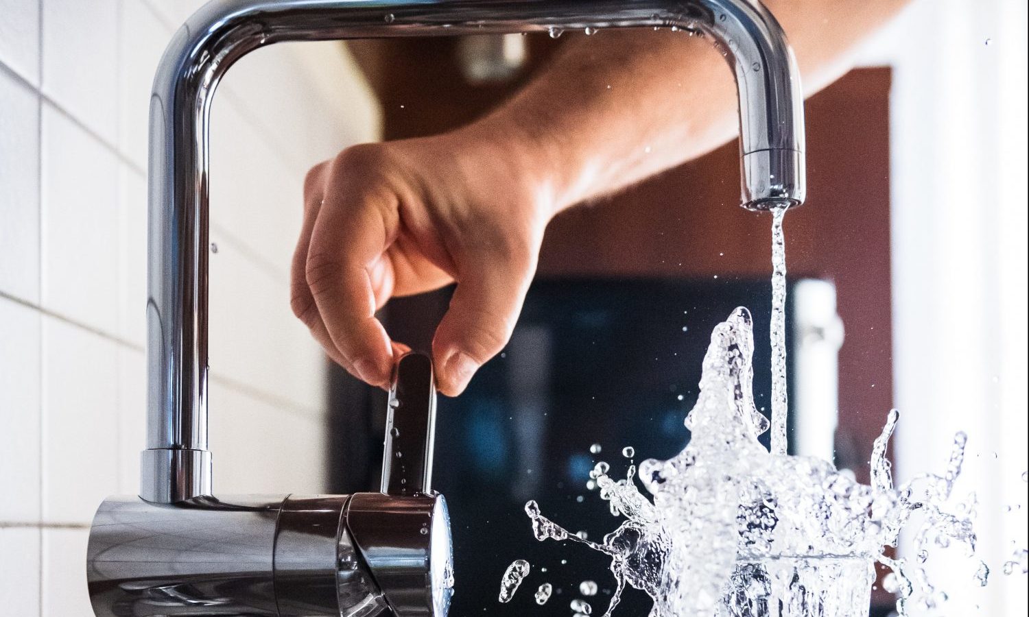 A white person's hand turning on a tap and filling up a glass of water, with the water splashing out of the glass.