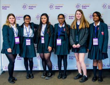 6 young people in school uniform are stood in front of a Longitude Explorer banner, smiling at the camera