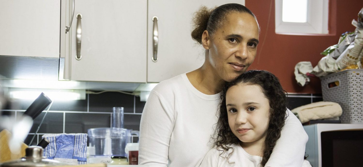 A woman wearing a white top stands with her arms around a young girl. They are standing in a kitchen, with grey tiles and white cupboards.
