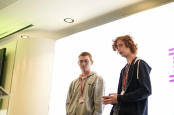 Two teenagers, both wearing hoodies, stand in front of a screen, presenting
