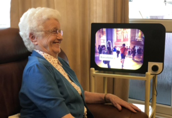 A smiling older woman sat in a chair, with a television next to her