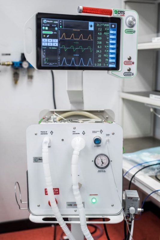 Black ventilator screen used in Chile during the COVID pandemic with various medical stats in different colours - looks like an EKG - Orange horizontal tube at the top right - white hospital wall background. PPE made in chile