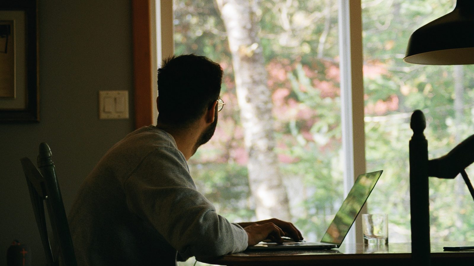 Man in grey T-shirt sat at a table with a laptop, staring out the window