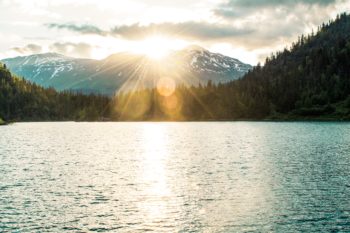 The sun sets behind the peaks of a mountain range in the distance. In front of it, fir trees line the lake that makes up most of the image.