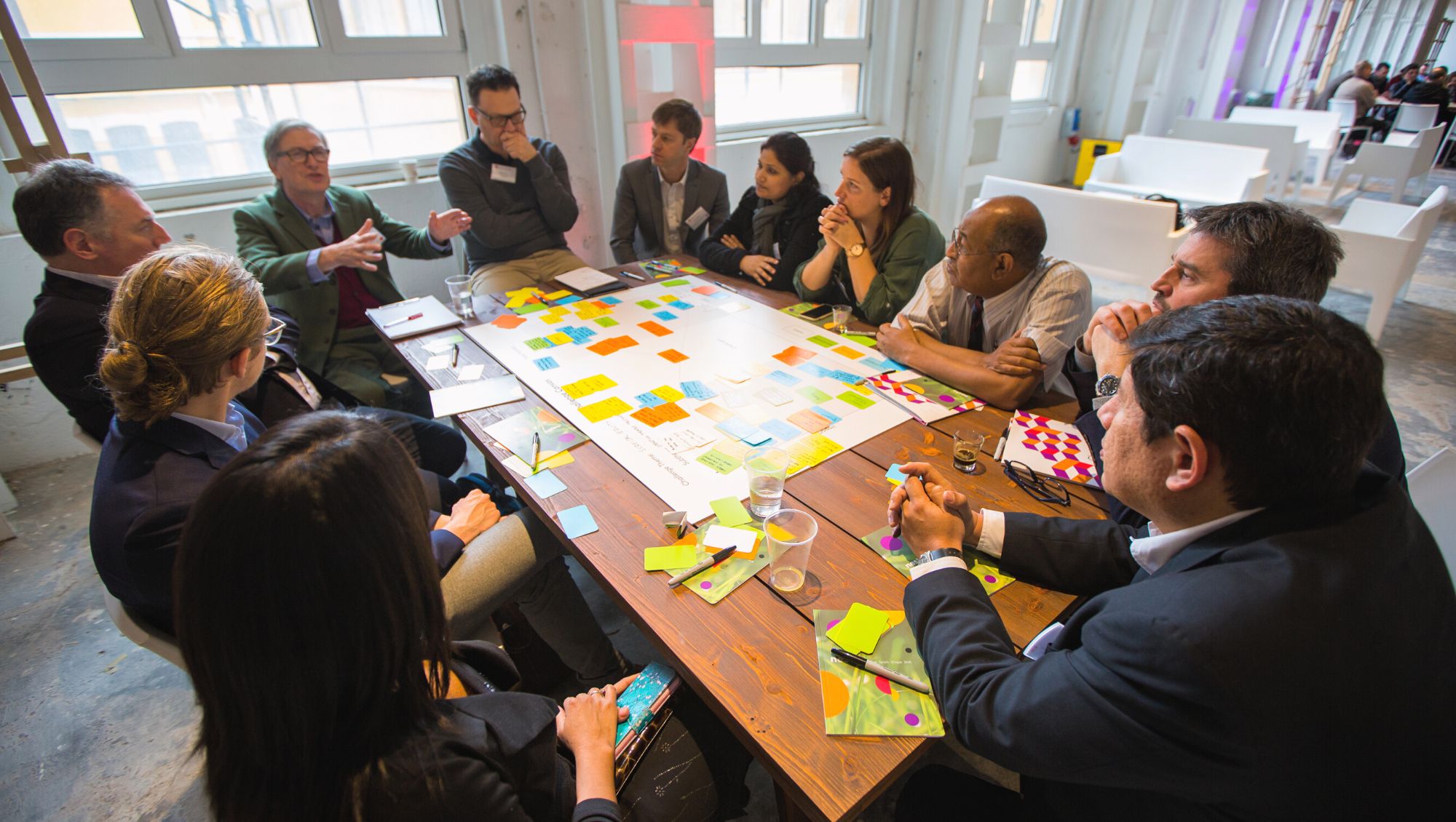 A group in business and smart casual clothes sit talking around a table strewn with ideas filled post-it notes