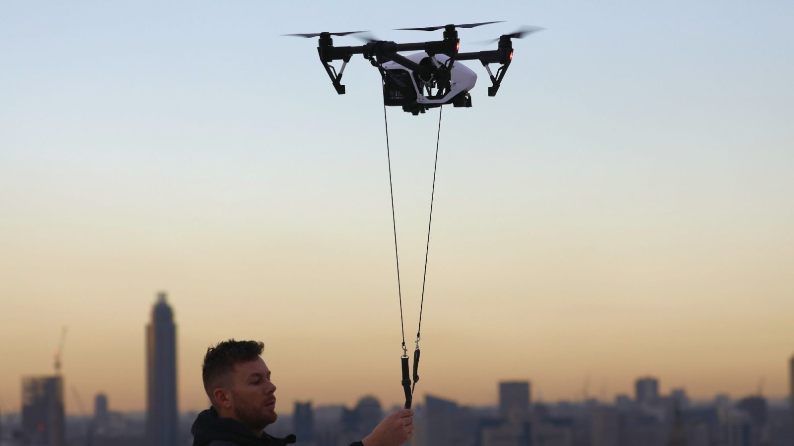 Man holds a hovering drone on a string in an open space in front of a the city skyline