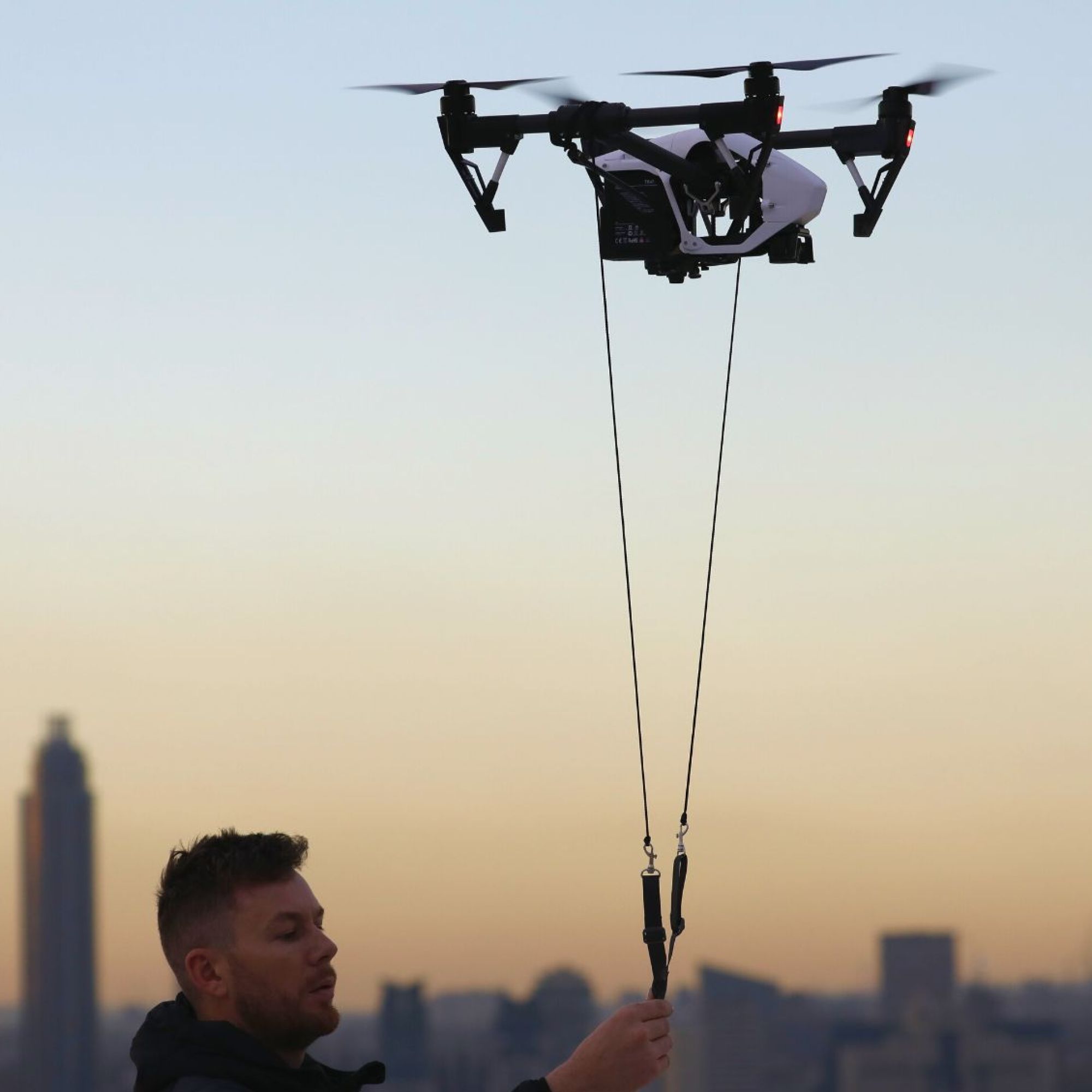 Man holds a hovering drone on a string in an open space in front of a the city skyline