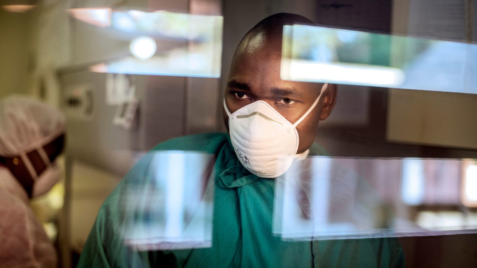 Man in green surgical scrubs and mask looks through a window with reflections, behind him a woman with mask and PPE works in the background