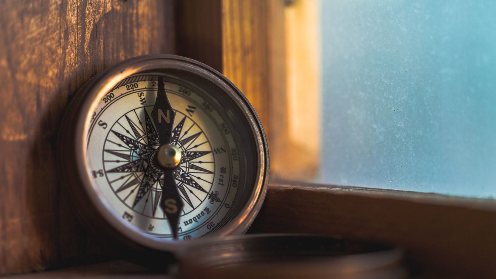 Antique compass rests open on a wooden sill next to a window representing the history of the Longitude Committee