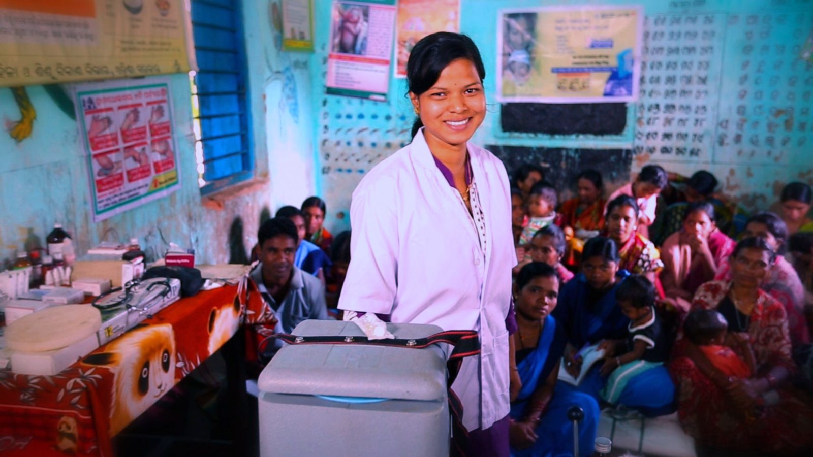 Woman in white coat stands smiling next to an insulated box in what appears to be a medical centre of sorts. Behind her a group of adults, some with babies and children sit on cushions the floor behind her wearing colourful saris.