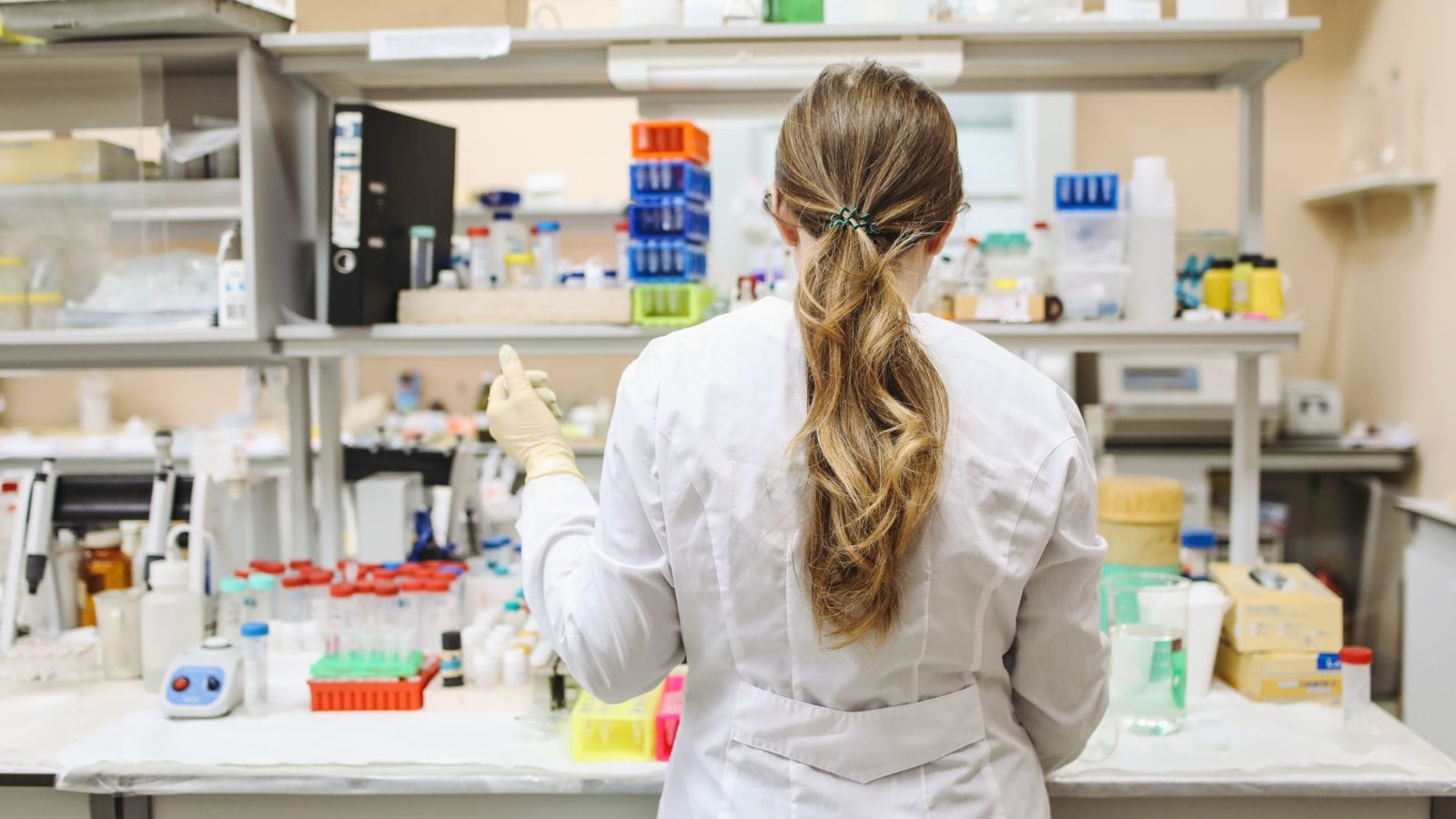 Facing away from camera a medical worker or biologist, wearing white coat and gloves, works at a medicine station filled with vials and equipment
