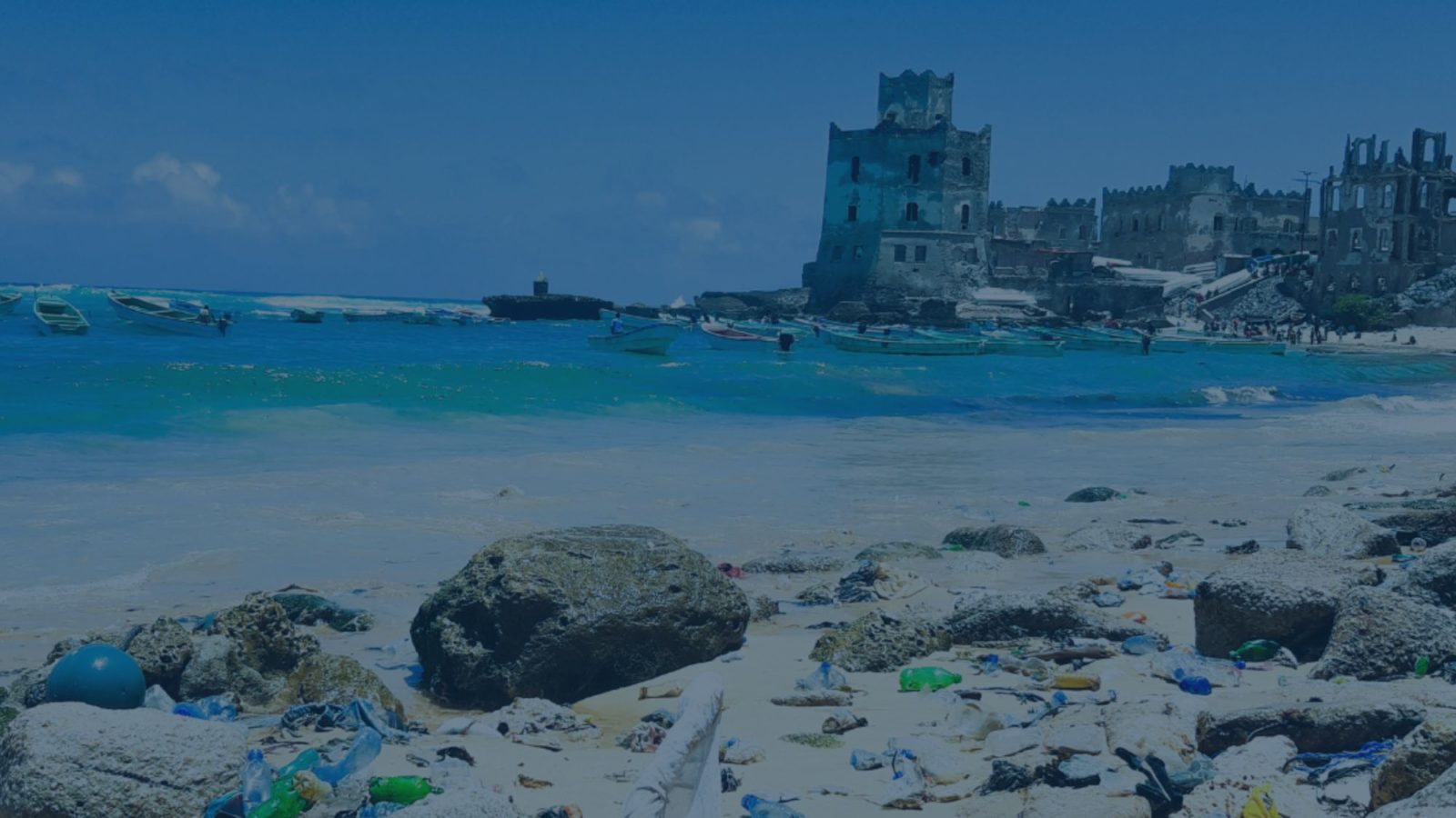 Plastic waste on a beach with rowing boats moored slightly out to sea and the ruins of a castle fort in the background