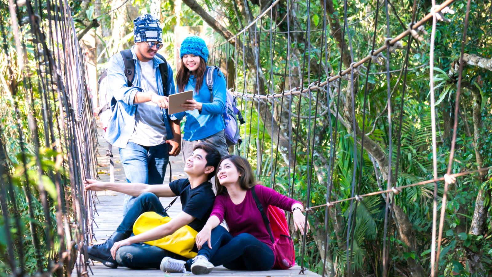 Four young tourists out on a sunny day, two looking at a tablet or notebook two sat on the bridge looking up. They are surrounded by lush green vegetation