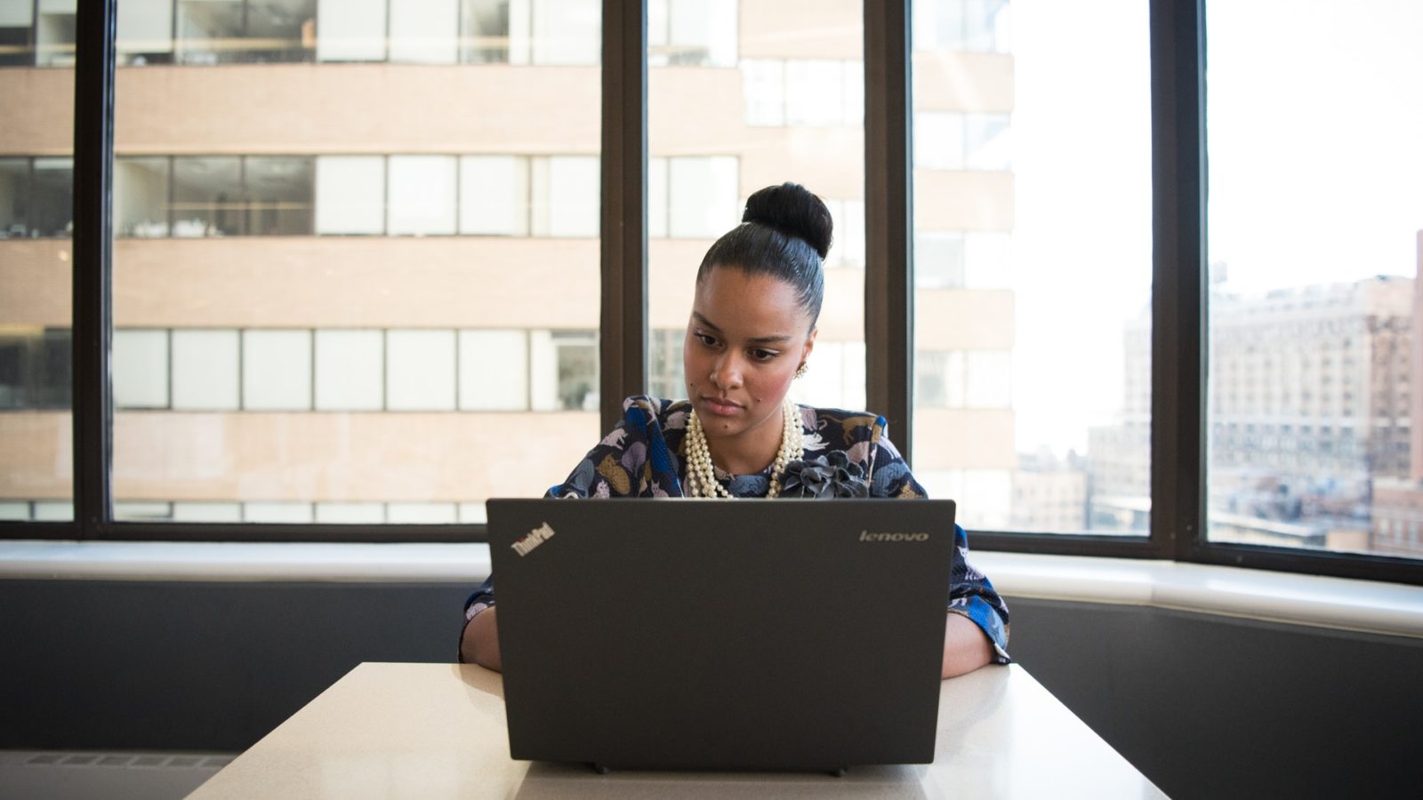 Woman with her hair in a high bun, sitting at a desk in front of wide windows, working on a laptop