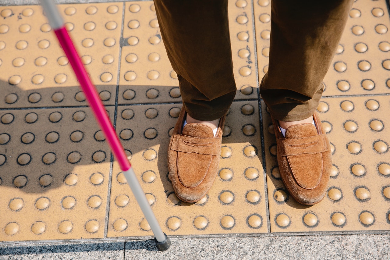 Looking down at legs at a pedestrian road crossing. Corduroy trousers and slippers with a deaf blind cane