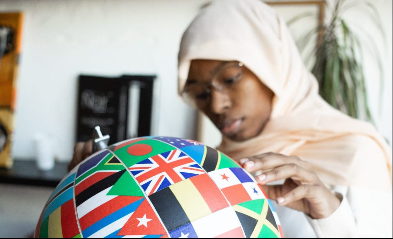 A woman wearing a hijab wearing glasses and touching a globe made with world flags
