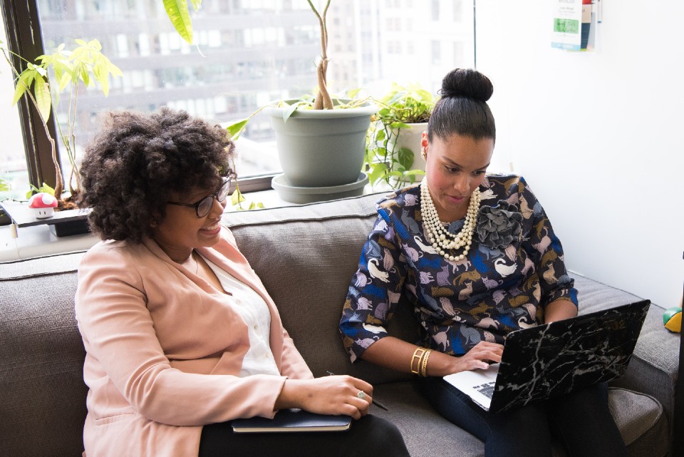 Two women sit on a couch looking at a laptop