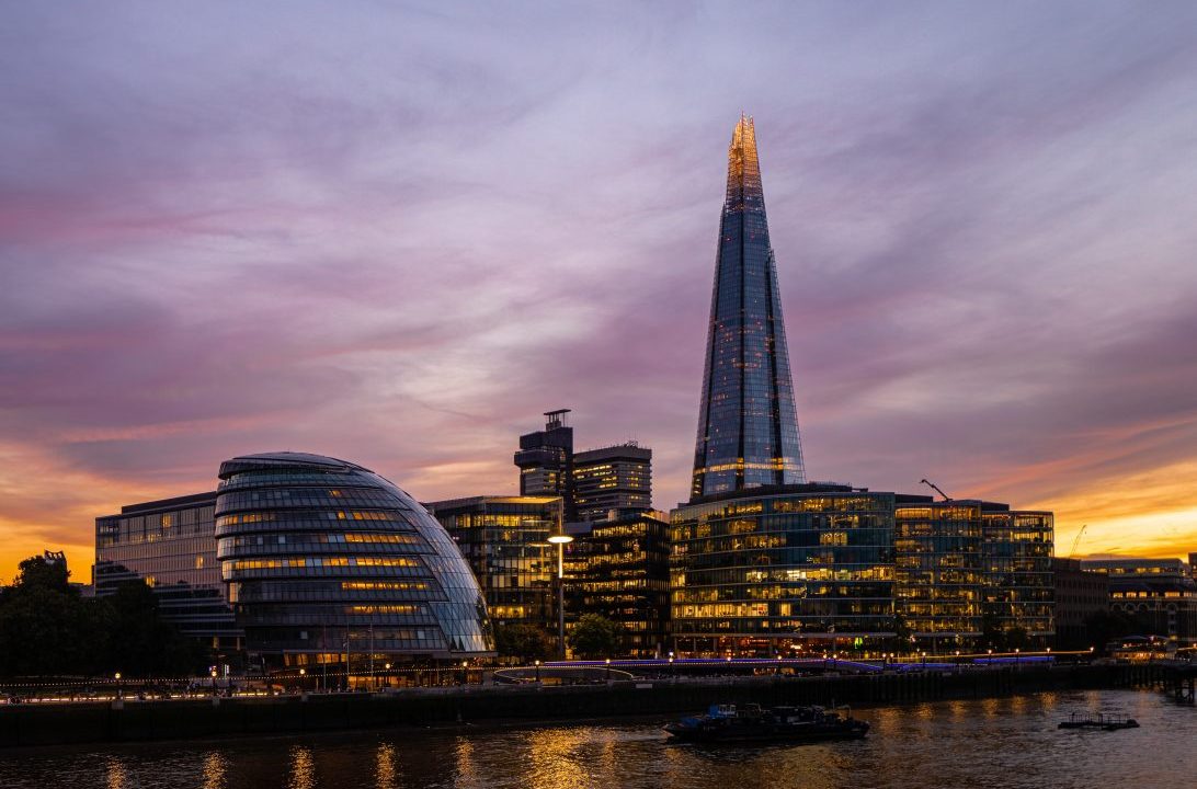A view of city hall and the shard on the River Thames at dusk with an orange light from the sun behind and on the river and light purple clouds