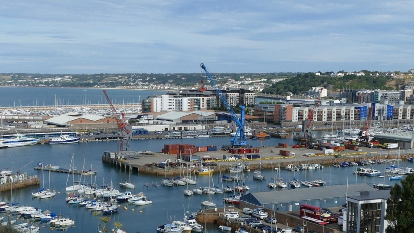 View of boats moored at the Port of St Helier, overlooked by modern buildings, with the island coast stretching into the distance