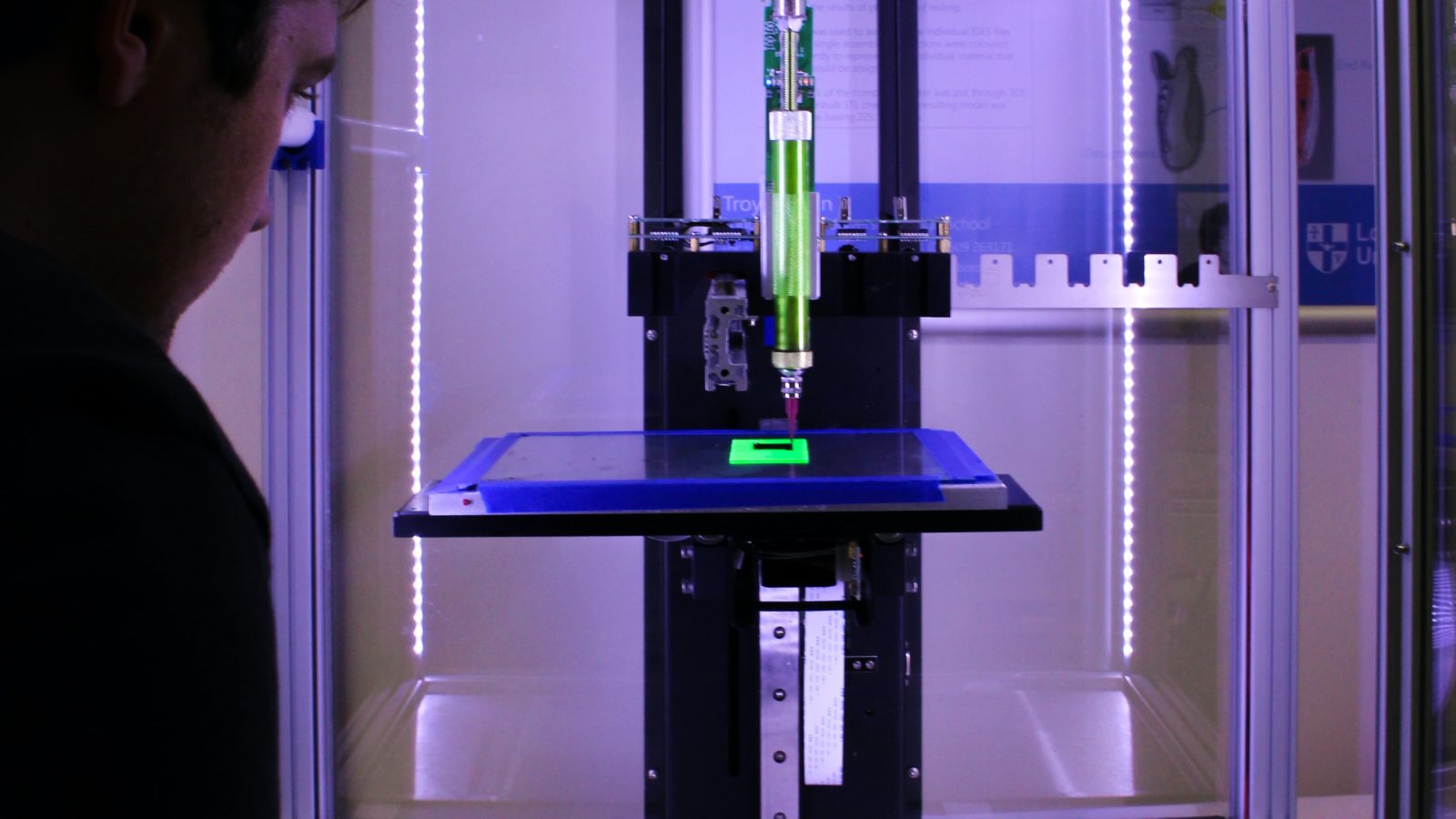 3d printer in a purple light with a person with white skin and short hair in shadows onlooking