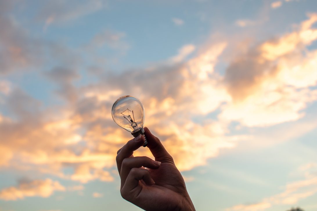 A hand holds a lightbulb up against a cloudy sky