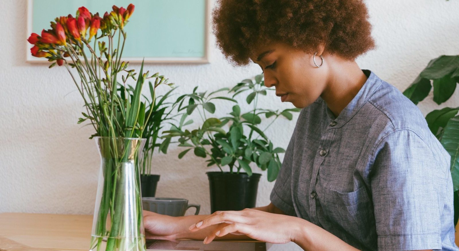 Someone who presents as female sits at a table browsing an iPad. There are flowers in a vase on the table.