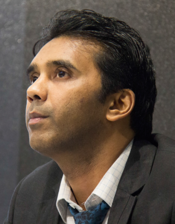 An Indian man wearing a dark suit and a white shirt, looks off to the left of the camera