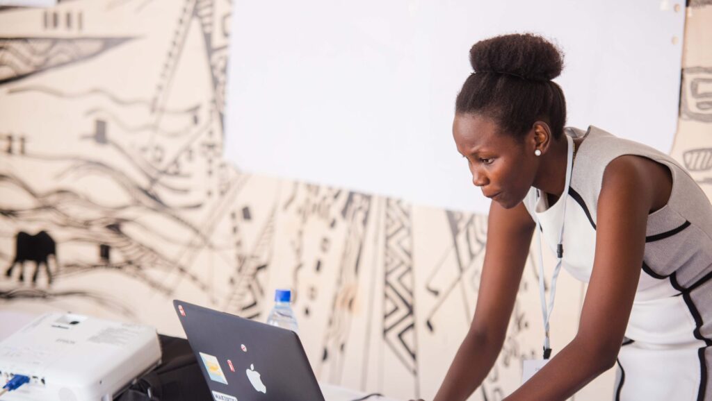 Black woman in a white dress leans over her laptop|CareerEar Founders Claudine Adyemi and Precious Ene|The founders of the Fall Army Worm Tech Challenge stand at a podium table and face the camera|