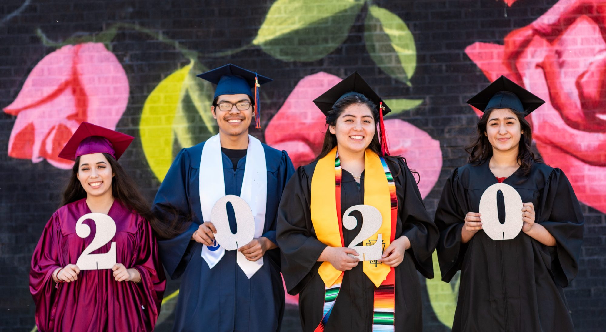 Four graduates stand in different colour robes holding the numbers for 2020. There is a floral pattern behind them with big pink roses.|An image of some cancer cells. They have been brightly coloured.|High angle shot of an unrecognizable businessman banking on his mobile phone|Three Tech to Connect trophies in a row|People walking under a bridge on a busy urban street|Surgical instruments in an operating theatre during cardiac surgery.|A group of school children pose for the Longitude Explorer Prize|A young man in a mask poses alongside his laptop|Open banking brand image: A line-drawn abstract femme looking person with red trousers