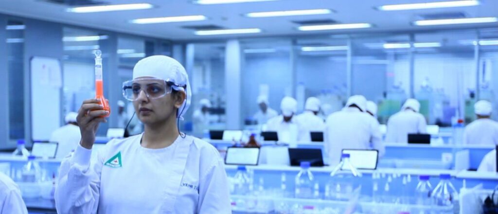 A person who presents as a woman in a research lab stands to the left holding a test tube filled with orange liquid. She is wearing a white lab coat and lap cap.|Pranav Johri from India stands smiling at the camera in a black suit jacket and white shirt - he has recovered from antibiotic resistance with phage therapy