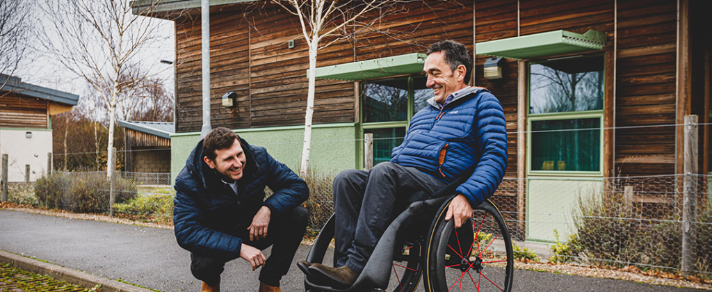 Two men on a path in front of a wooden building. One is crouched down next to the other