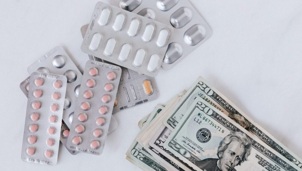 Silver blister packets of various pills next to a stack of American bank notes|Mary Millard