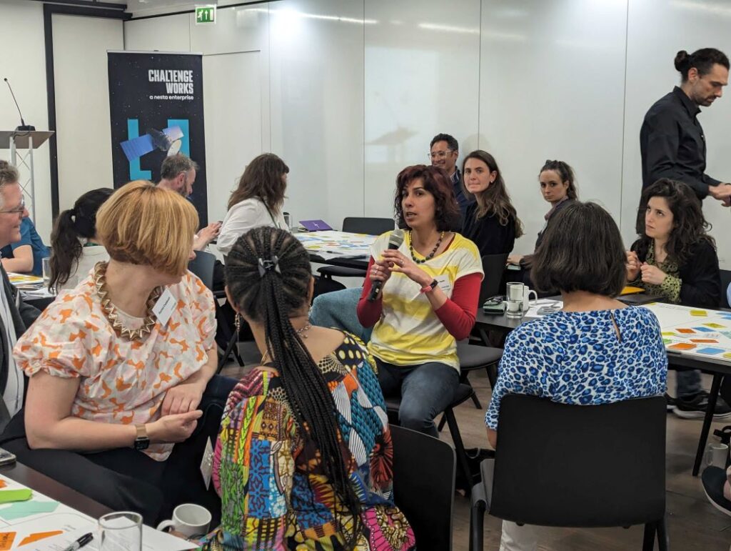A group of people attending a challenge works event. They are sitting in chairs crowding around a women who is speaking on a mic.|Groups of people at a challenge works event sitting in tables discussing idea amongst each other.|A group of people attending a challenge works event. They are sitting in chairs crowding around a women who is speaking on a mic.