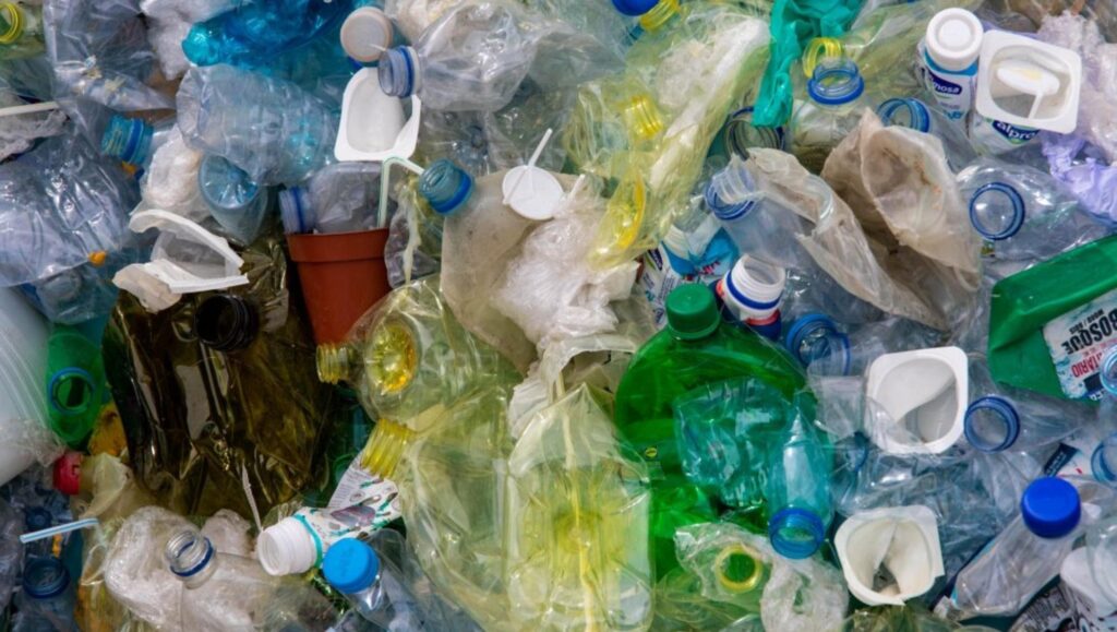 Plastic waste|A hand holding a phone and using it to scan barcodes|A blue spray bottle with orange writing on it saying 'spray safe'|A group of people stand outside in front of a large sign saying VEnvirotech