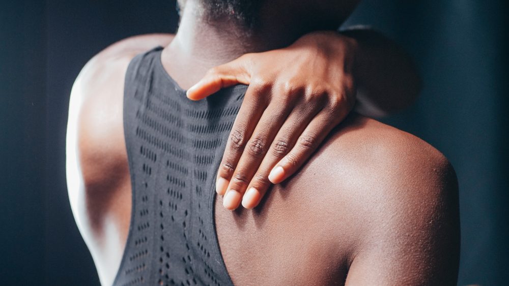 A person with dark skin wearing a black razor back tank is facing away from us and has their hand on their shoulder like they are in pain