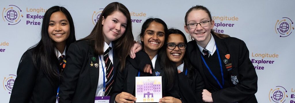 A group of five teenagers in school uniform smile at the camera while standing in front of a post for the Longitude Explorer Prize|A purple and yellow illustration titled Ocean Cleanup shows a sailboat