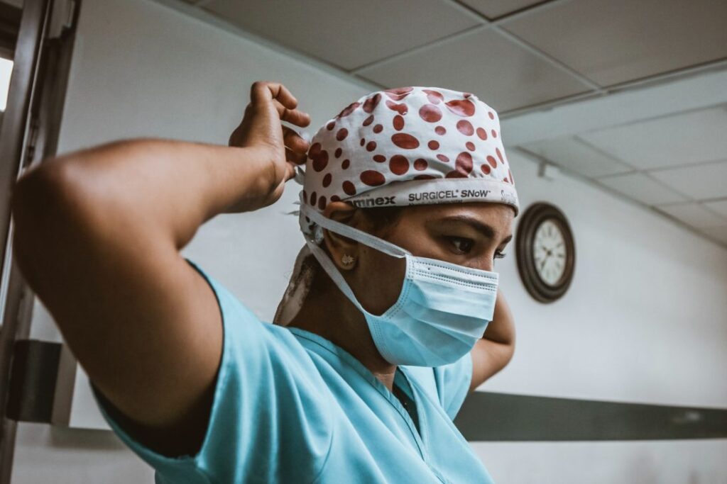 An Indian surgeon in teal scrubs and a patterned headscarf is securing a face mask in a white corridor|An Indian man wearing a dark suit and a white shirt