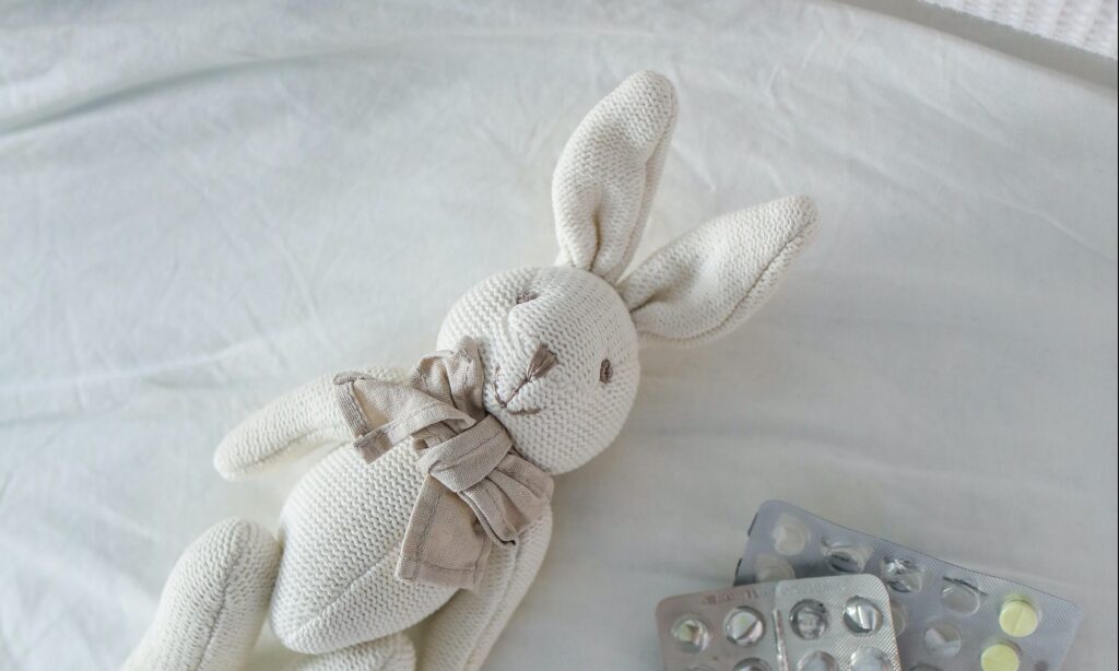 A white toy stuffed bunny lays on a bed next to some medicine packets that have some pills missing