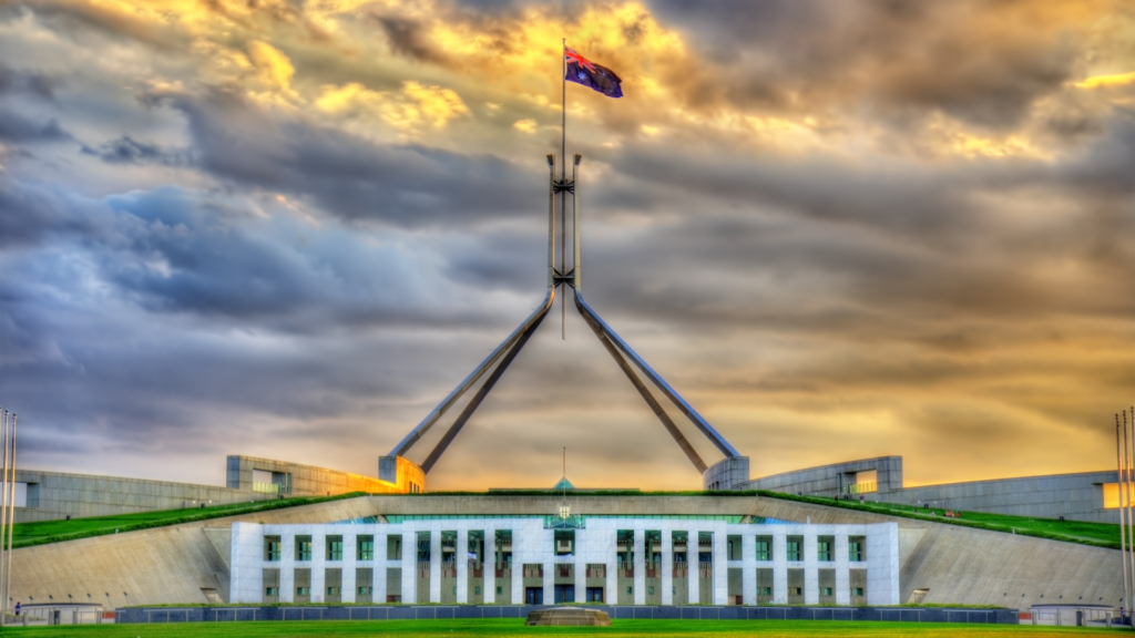 A photo of the Australian Parliament building at dawn. A cloudy sky is lit up behind an Australian flag.|A photo of the Australian Parliament building at dawn. A cloudy sky is lit up behind an Australian flag.