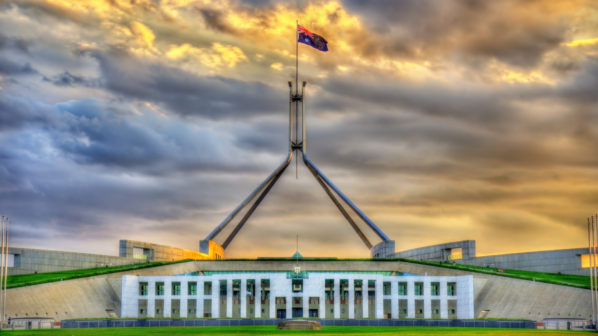 A photo of the Australian Parliament building at dawn. A cloudy sky is lit up behind an Australian flag.|A photo of the Australian Parliament building at dawn. A cloudy sky is lit up behind an Australian flag.