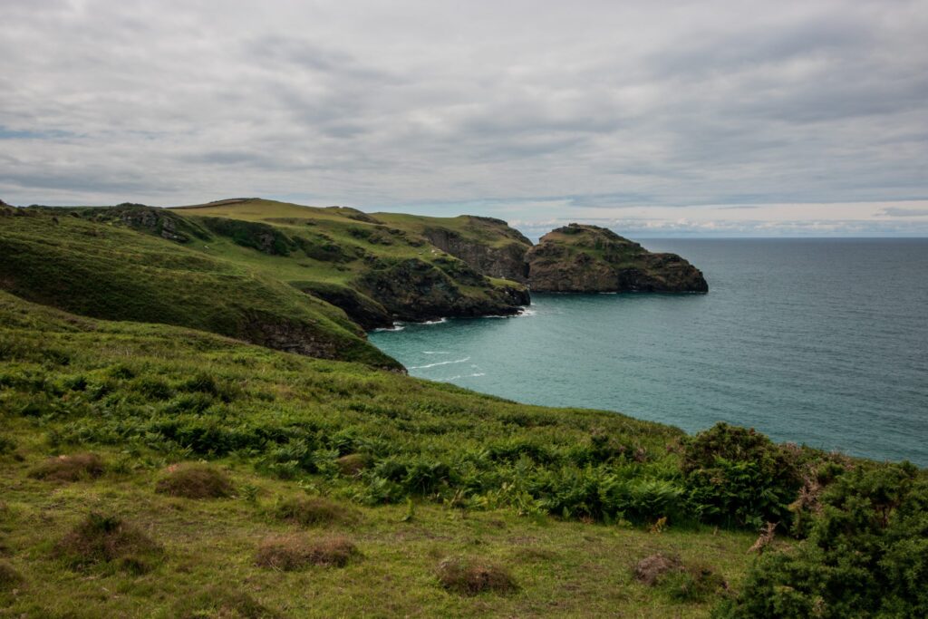 A lush green spit of coast curves around as it meets the sea|The sun sets behind the peaks of a mountain range in the distance. In front of it