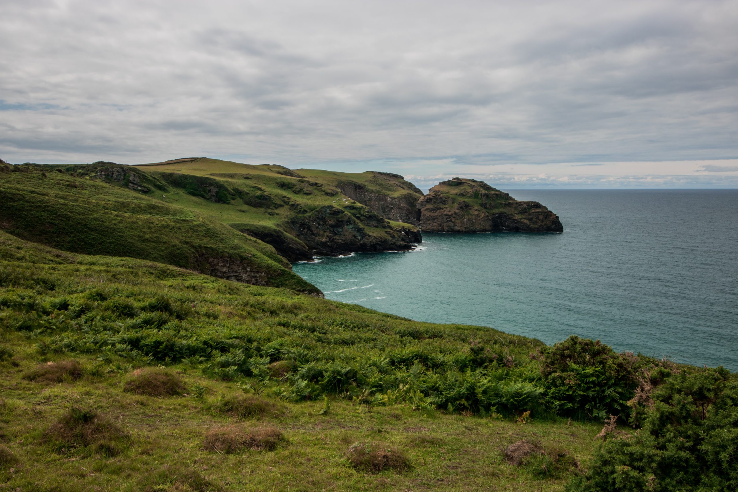 A lush green spit of coast curves around as it meets the sea|The sun sets behind the peaks of a mountain range in the distance. In front of it