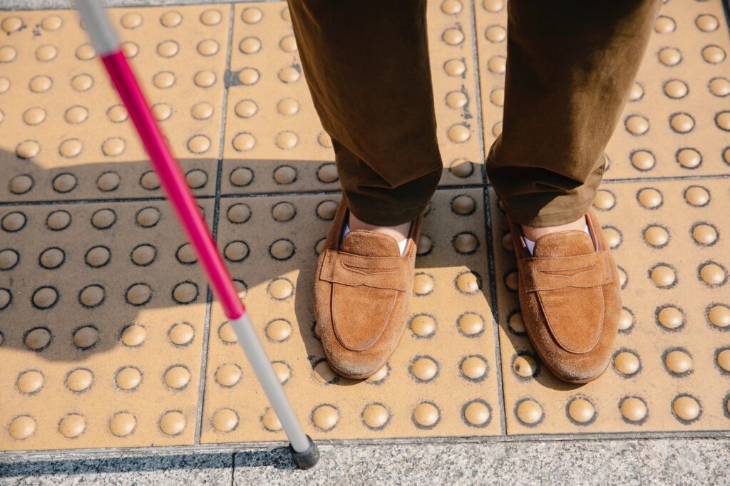 Looking down at legs at a pedestrian road crossing. Corduroy trousers and slippers with a deaf blind cane|Horizon 2020 logo. European Union funding for Research & Innovation.